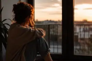 Vrouw zit alleen bij het raam bij zonsondergang als beeld voor de pijn van onveilige hechting in relaties