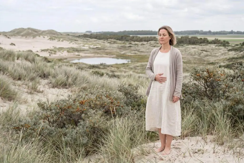 Vrouw staat rustig in de duinen met een hand op haar buik en gesloten ogen, beeld bij stoppen met pleasen