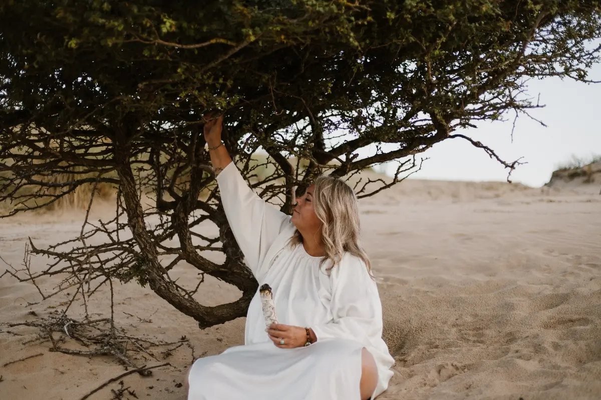 Helen Adriana zit alleen onder een windgevormde boom in de duinen, in een witte jurk, met haar hand omhoog naar de takken